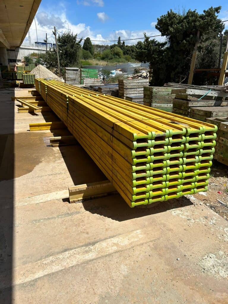 ערימה ארוכה של קורות עץ צהובות עם קצוות ירוקים, מונחת על הקרקע