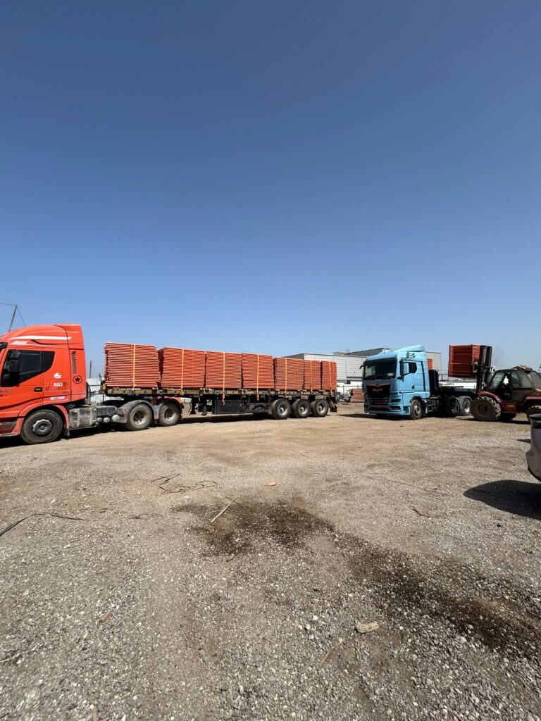 משאית עם קבינה אדומה ונגרר עמוס במסגרות מתכת כתומות